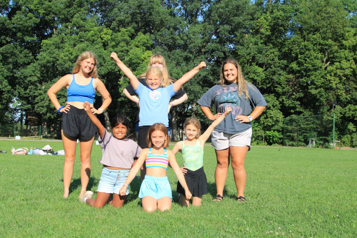 A group of girls doing cheerleading poses outdoors with their camp counselors.